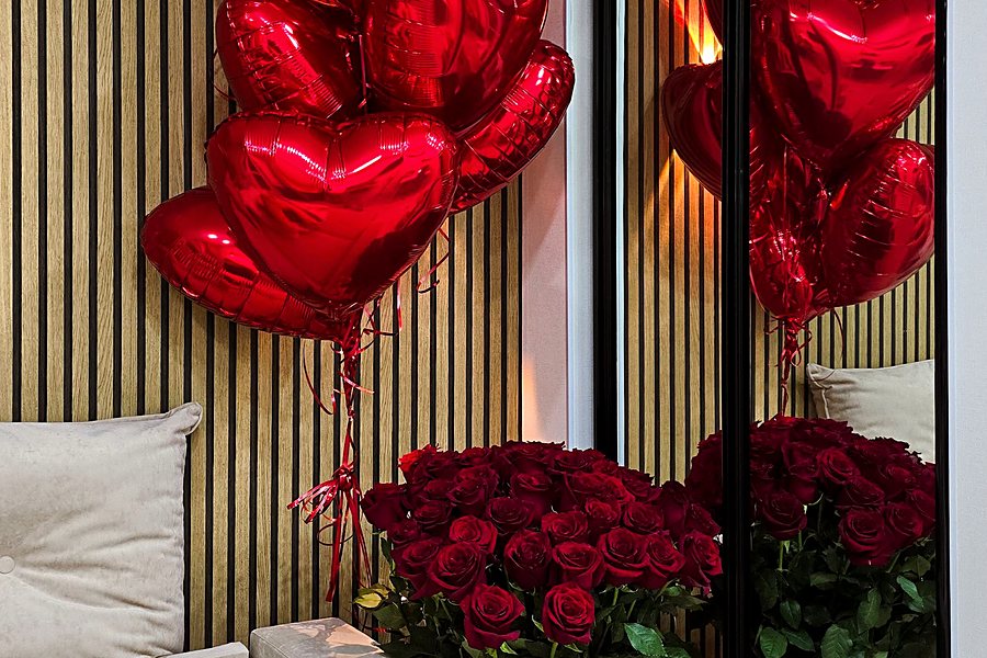 Room styled with red heart balloons, a large arrangement of red roses and tulips beside a sofa