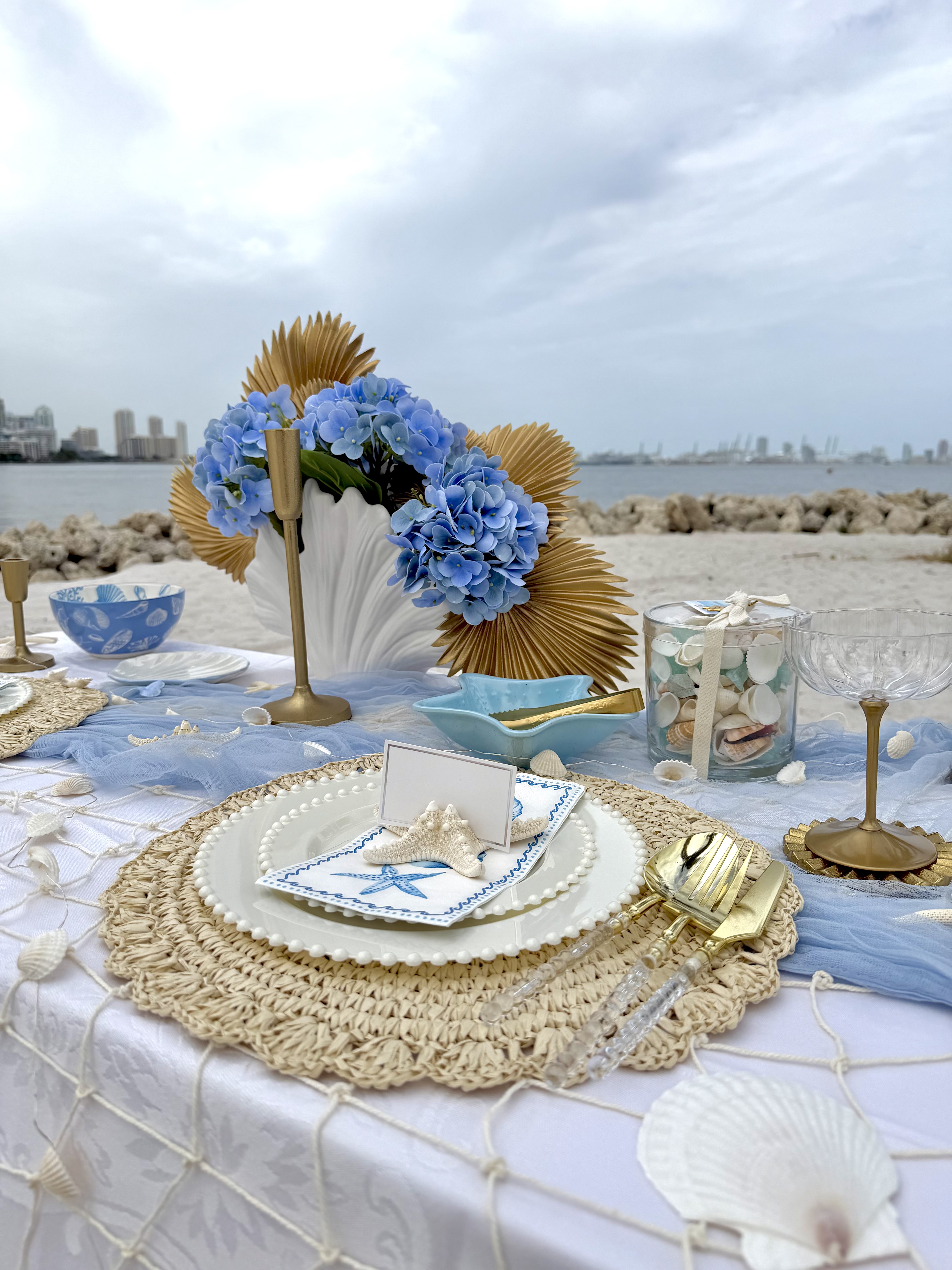 Beach table setting with blue hydrangeas, gold candlesticks, shells and a woven charger by the water